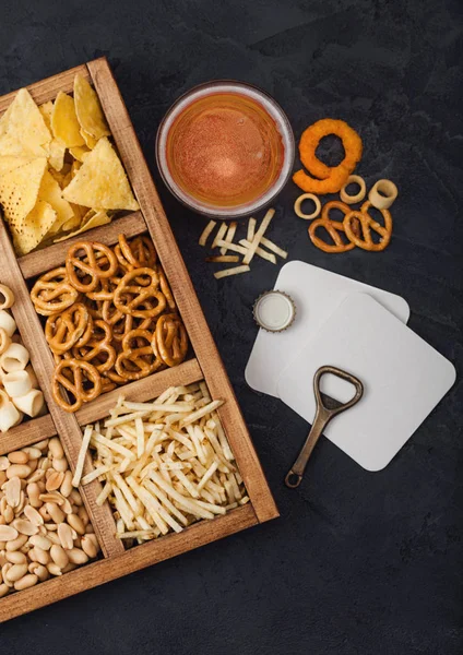 Vaso de cerveza artesanal y abrelatas con caja de snacks sobre fondo negro. Pretzel, palitos de patata salada, cacahuetes, aros de cebolla con nachos en caja vintage con abrelatas y esterillas de cerveza. Macro — Foto de Stock