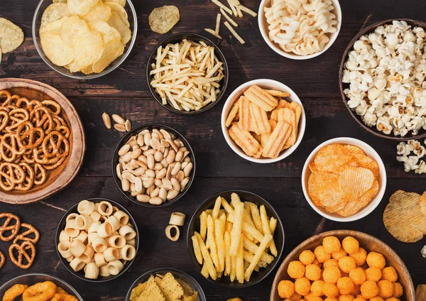 All classic potato snacks with peanuts, popcorn and onion rings and salted pretzels in bowl plates on wooden background. Twirls with sticks and potato chips and crisps with nachos and cheese balls. — Stock Photo, Image