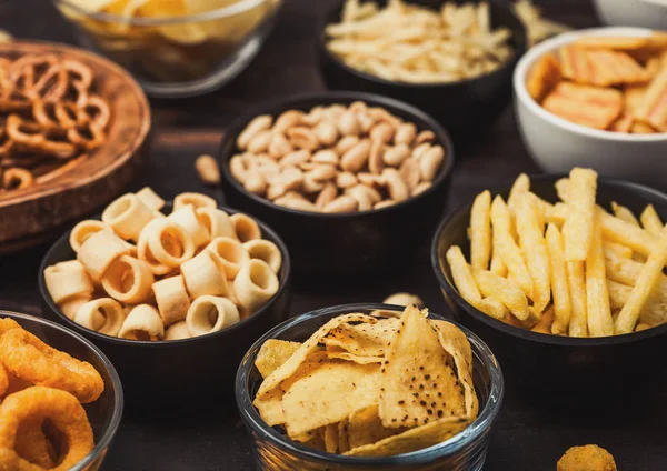 Todos los snacks clásicos de papa con cacahuetes, palomitas de maíz y aros de cebolla y pretzels salados en platos hondos sobre fondo de madera. Giros con palitos y papas fritas y patatas fritas con nachos y bolas de queso . — Foto de Stock