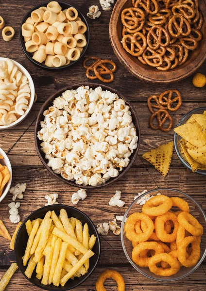 All classic potato snacks with peanuts, popcorn and onion rings and salted pretzels in bowl plates on wooden background. Twirls with sticks and potato chips and crisps with nachos and cheese balls. — Stock Photo, Image