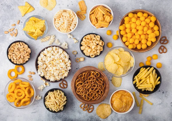 Todos los snacks clásicos de papa con cacahuetes, palomitas de maíz y aros de cebolla y pretzels salados en platos hondos sobre fondo claro. Giros con palitos y papas fritas y patatas fritas con nachos y bolas de queso . — Foto de Stock