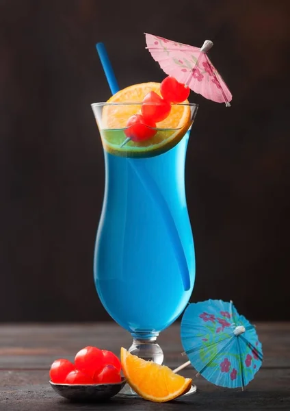 Blue lagoon summer cocktail in classic glass with sweet cocktail cherries and orange slice with umbrella on dark table background.
