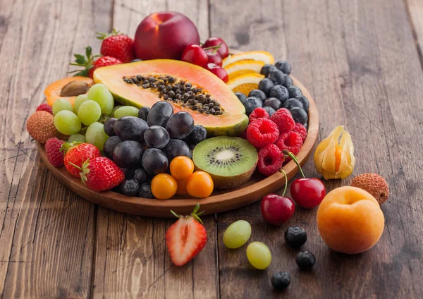 Taze çiğ organik yaz meyveleri ve ahşap mutfak arka planında yuvarlak ahşap tabakta egzotik meyveler. Papaya, üzüm, nektarin, portakal, ahududu, kivi, çilek, liçi, kiraz. Üst görünüm