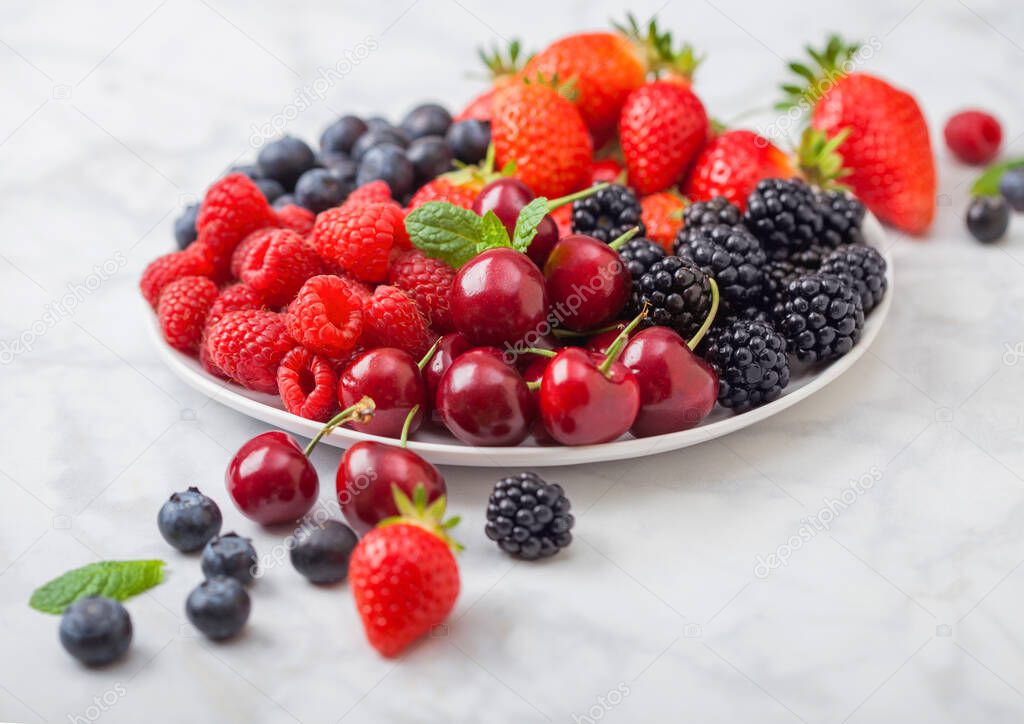 Fresh organic summer berries mix in white plate on marble background. Raspberries, strawberries, blueberries, blackberries and cherries. Top view