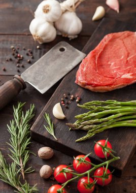 Beef braising steak, fresh raw slice on chopping board with garlic, asparagus and tomatoes with salt and pepper on wooden background with meat hatchet. clipart