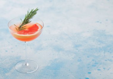 Kırmızı greyfurt yaz suyu kokteyli. Modern lüks martini bardağında. Biberiye ve açık mavi arka planda meyve dilimi. Metin için boşluk