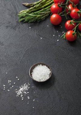 Siyah yemek arka planı kuşkonmaz, kiraz domatesleri ve siyah masa zemininde bir kase tuz. Dikey