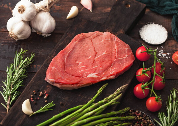 Raw fresh beef braising steak on chopping board with garlic, asparagus and tomatoes with salt and pepper on wooden table background. Macro