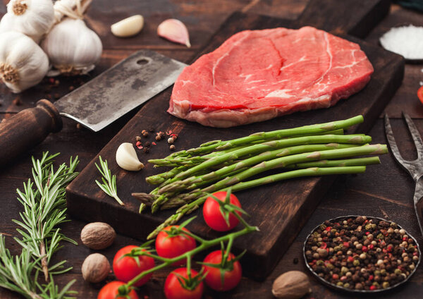 Slice of fresh raw barbeque braising beef steak on chopping board with asparagus and garlic with tomatoes and salt with pepper on wooden background with hatchet.