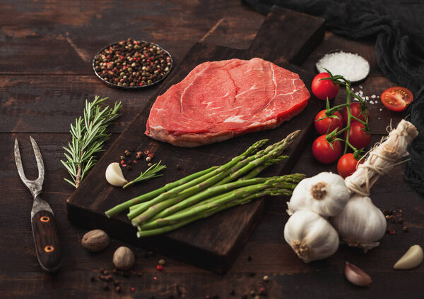 Slice of raw fresh beef braising steak on chopping board with garlic, asparagus and tomatoes with salt and pepper with rosemary and meat fork on wooden background.