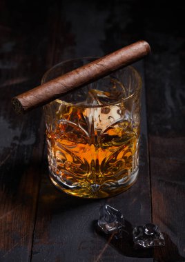 Tek malt viski, kristal bardaklarda buz küpleri ve tahta masa arkasında Küba purosu..