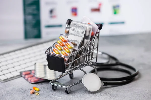 Shopping trolley with medicines for online purchase with keyboard and stethoscope on light background. Vitamins and antibiotics pills with painkillers and flu and cold capsules.