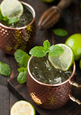 Bakır bardakta Moskova katırı kokteyli, kireç, nane ve ahşap sıkacağı koyu ahşap masa arkasında..