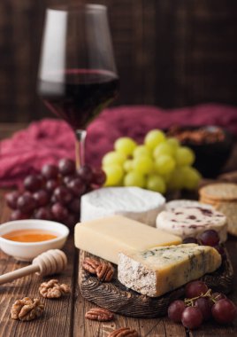 Ahşap masa arkasında çeşitli peynir ve üzümlerden oluşan bir bardak kırmızı şarap. Mavi Stilton, Kırmızı Leicester ve Brie Cheese ve bıçaklı ballı.