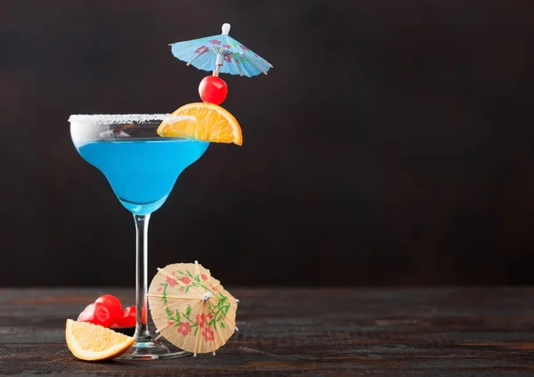 Blue lagoon summer cocktail in margarita glass with sweet cocktail cherries and orange slice with umbrella on dark table background. Space for text