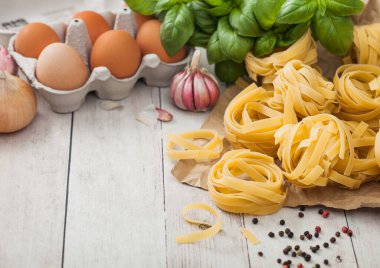 Beyaz arka planda kahverengi kağıtta ev yapımı tagliatelle makarna. Arka planda yumurta, fesleğen ve sarımsak var..