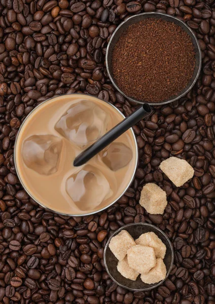 Glass of cold coffee with ice cubes and straw on fresh raw coffee beans background with cane sugar and powder. Top view