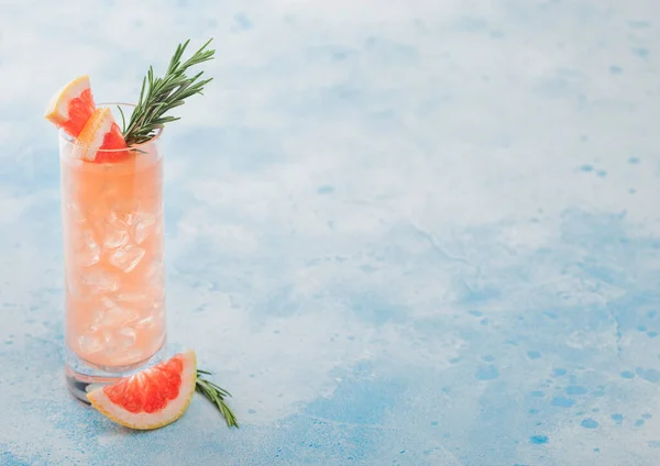 Highball glass of refreshing summer red grapefruit cocktail with ice cubes, fruit slice and rosemary on blue background. Space for text
