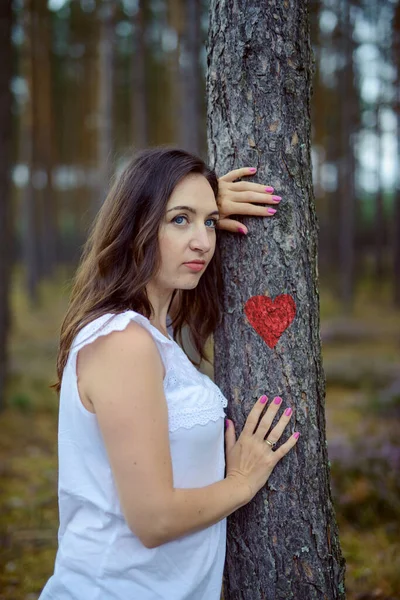 Mujer joven toma de la mano un árbol en el que se pinta un corazón rojo. amor por la naturaleza. espacio de copia. imagen auténtica . —  Fotos de Stock