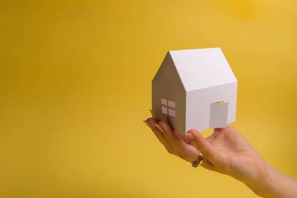 White family paper house in man hand on yellow background paper. Minimalistic style. Copy space. View from above. Vertical orientation