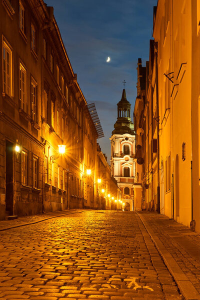 A cobbled street with a baroque belfry of a historic monastery at night in Poznan
