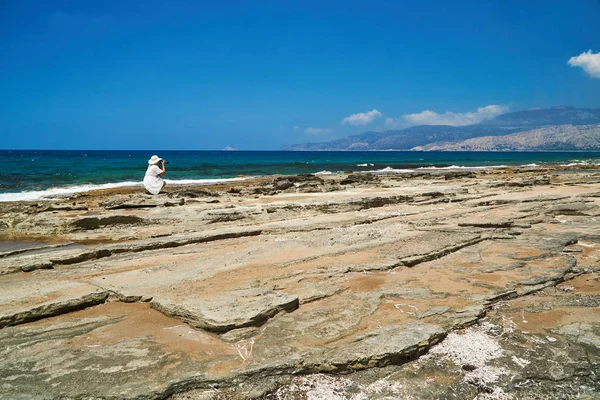 在罗得岛的岩石海滩上 穿着白色连衣裙和帽子的女孩 带着相机 — 图库照片