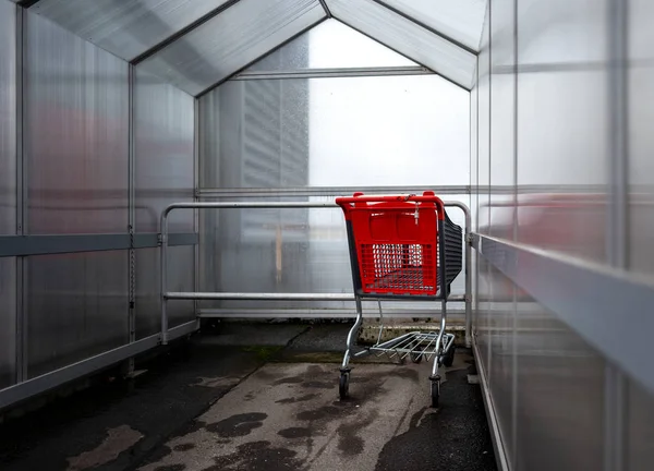 Último carrito de compras rojo bajo una cubierta de lluvia en frente de la entrada de una tienda —  Fotos de Stock
