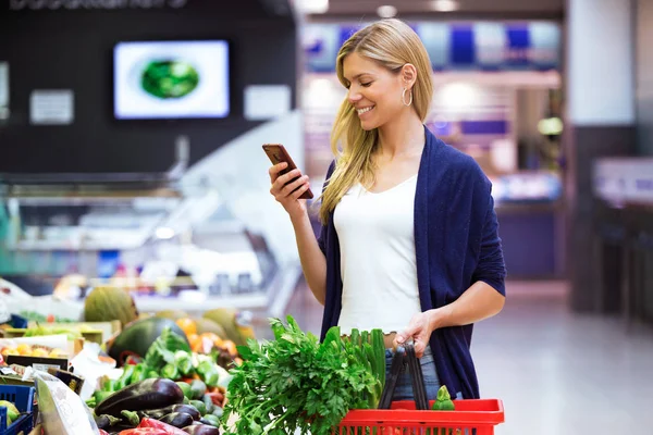 在市场上买新鲜蔬菜时 一张漂亮的年轻女子在手机上看购物清单 — 图库照片
