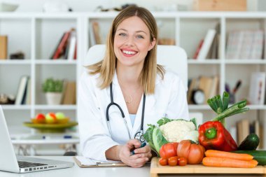 Portrait of beautiful smiling nutritionist looking at camera and showing healthy vegetables in the consultation. clipart