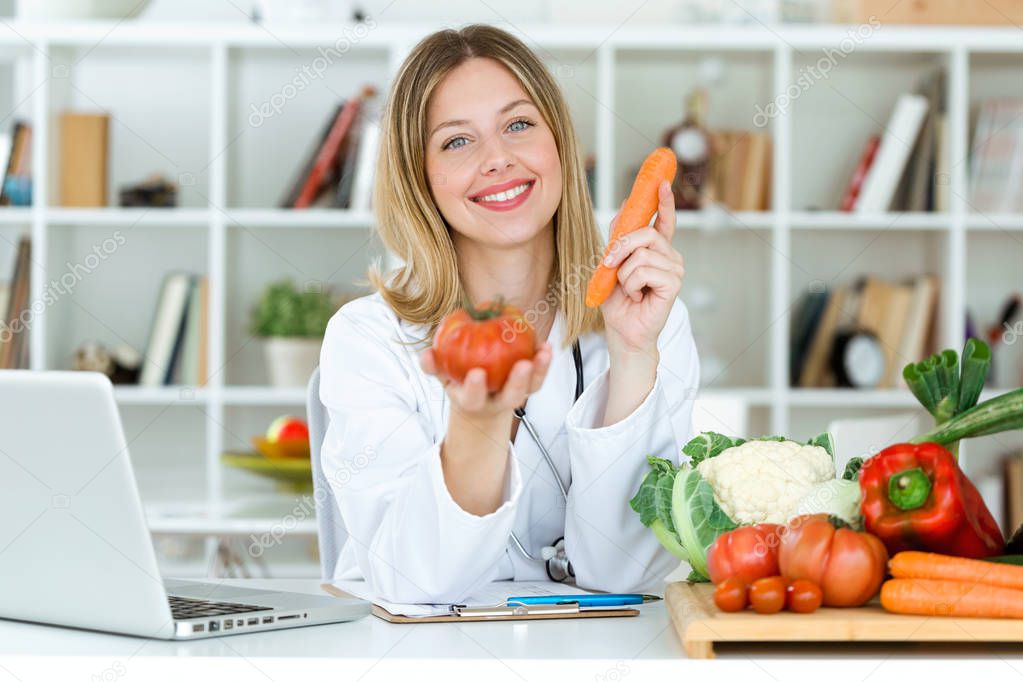 Portrait of beautiful young nutritionist looking at camera and holding fresh vegetables in the consultation.