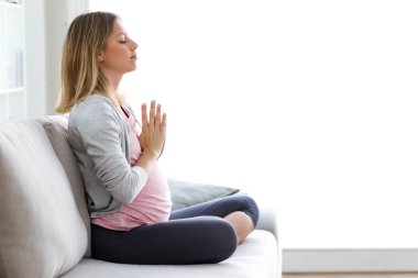 Shot of beautiful pregnant woman doing yoga, sitting in lotus position at home clipart