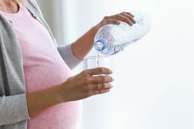 Close-up of pregnant young woman putting water in a glass at home clipart