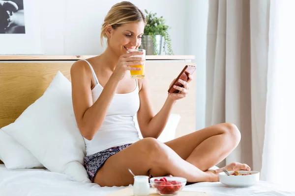 寝室での朝食と彼女の携帯電話にテキスト メッセージを持つ美しい若い女性のショット — ストック写真