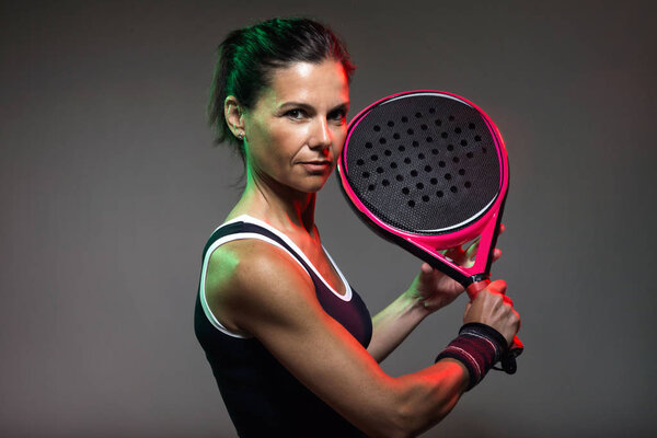 Portrait of adult fitness woman looking at camera while playing padel indoor. Isolated on black.