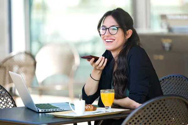 Vacker ung kvinna talar med sin mobiltelefon medan du äter frukost medan du arbetar med sin laptop på kafé. — Stockfoto