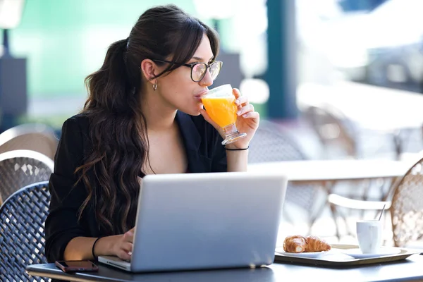 在咖啡店里用笔记本电脑工作时 年轻女子喝橙汁的镜头 — 图库照片