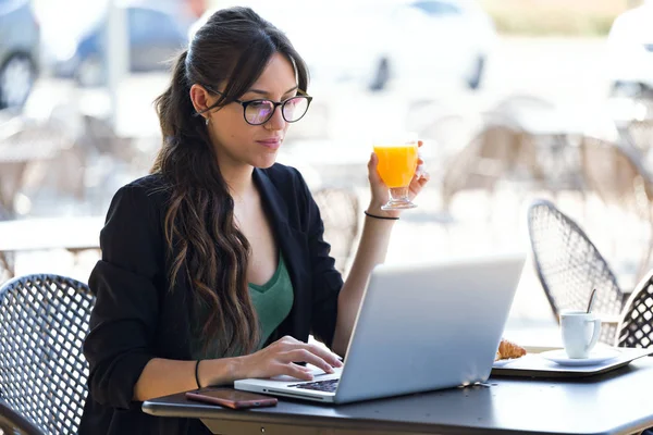 在咖啡店里用笔记本电脑工作时 年轻女子喝橙汁的镜头 — 图库照片