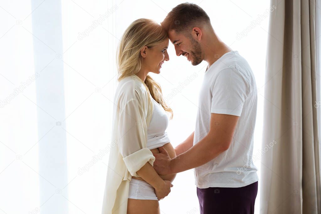 Shot of beautiful young couple expecting baby standing together and touching belly near the windows at home.