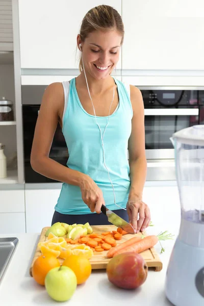 台所で音楽を聴きながらいくつかの野菜や果物を切る美しい若いスポーティな女性のショット — ストック写真