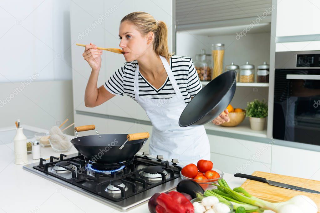 Shot of healthy young woman cooking and tasting food with wooden spoon in the kitchen at home. 