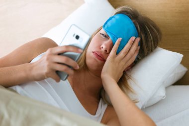 Ne zaman evde yatak odasında smartphone olduğunu görmek için uyku maskesi çekerek yatakta yalan yorgun genç kadının atış.