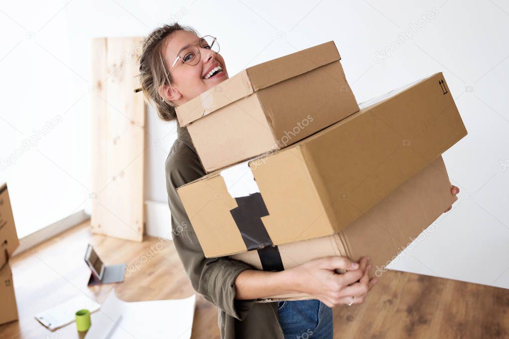 Shot of pretty young woman carrying cardboard boxes into her new house while smiling at camera.