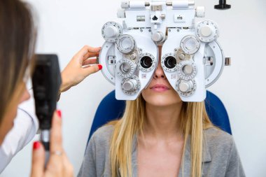Shot of young woman sitting on chair with beautiful optician standing while doing eye test in ophthalmology clinic. clipart