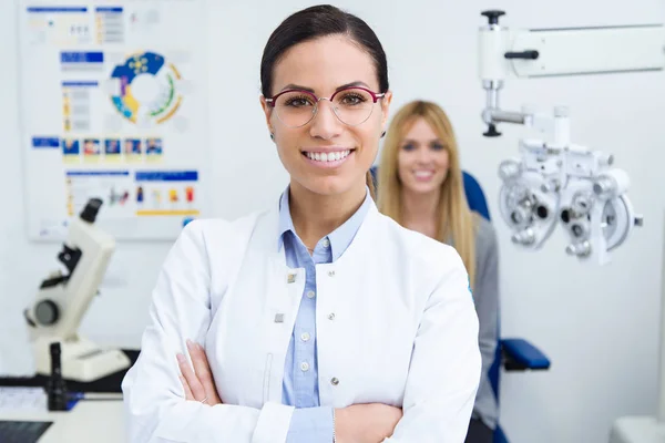 美丽微笑的妇女的肖像看着相机在眼科诊所与女性患者坐在眼睛测试的背景 — 图库照片