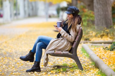 Güzel genç kadın bir Bank sokakta oturuyor ve kahve içme sırasında cep telefonu kullanarak çekim.
