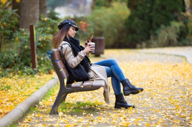 Güzel genç kadın bir Bank sokakta oturuyor ve kahve içme sırasında cep telefonu kullanarak çekim.