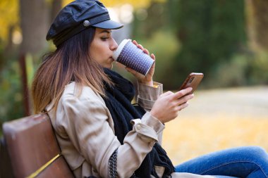 Güzel genç kadın bir Bank sokakta oturuyor ve kahve içme sırasında cep telefonu kullanarak çekim.