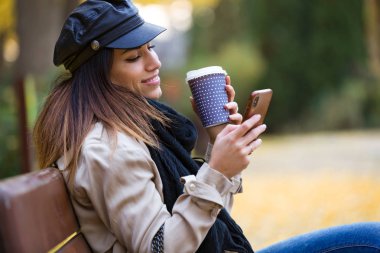 Güzel genç kadın bir Bank sokakta oturuyor ve kahve içme sırasında cep telefonu kullanarak çekim.