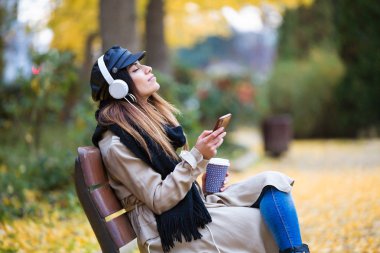 Genç ve güzel kadın parkta cep telefonu ile müzik dinlerken bir Bank rahatlatıcı kadeh.