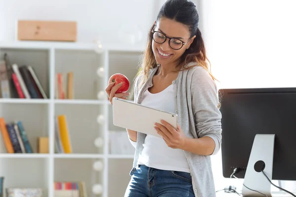 Mooie jonge zakenvrouw met behulp van haar digitale tablet terwijl het eten van rode appel in het kantoor. — Stockfoto